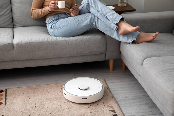 Modern robot vacuum cleaner near sofa with resting woman in room - Photo, Image