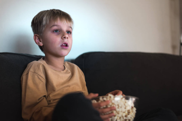 Цікава дитина їсть попкорн під час перегляду телевізора з цікавістю на дивані
 - Фото, зображення
