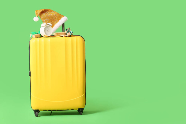 Желтый чемодан с рождественскими подарочными коробками, будильником, деревянным самолетом и шляпой Санты на зеленом фоне - Фото, изображение