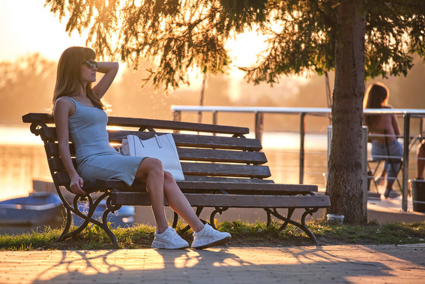 Samotna kobieta siedząca na ławce nad jeziorem ciesząca się ciepłym letnim wieczorem. Pojęcie samotności i relaksu - Zdjęcie, obraz