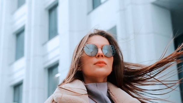 A girl in a beige jacket and glasses walks down the street in the business district of the city, a strong wind. Success - Фото, изображение