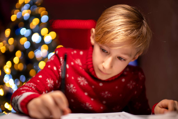 poika punaisessa joulupuserossa joulukuusen taustalla piirtää jotain huoneeseensa - Valokuva, kuva