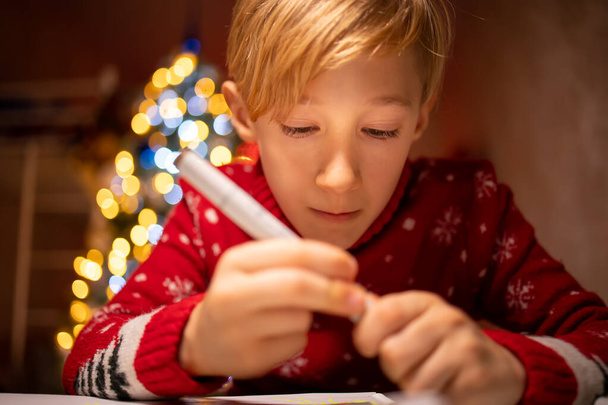 chlapec v červeném vánočním svetru na pozadí vánočního stromku se zabývá procesem kreslení ve svém pokoji pod lampou - Fotografie, Obrázek