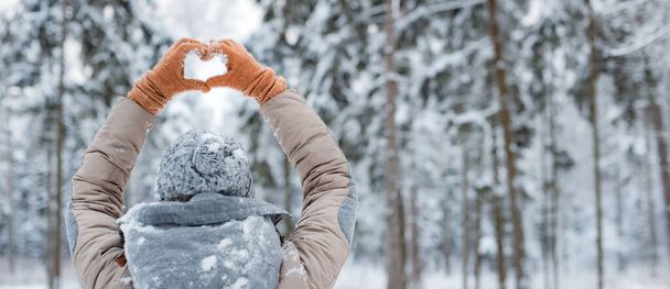 happy woman walking outside woods snowy winter day. woman mittens dressed fashionable gray stands with her back makes heart gesture with her hands against background snowy winter forest - Photo, Image