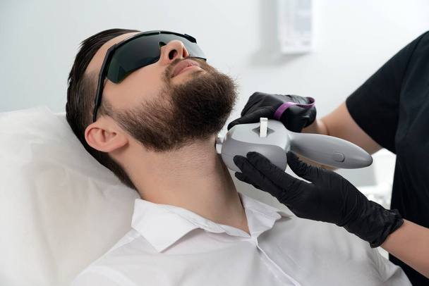 Молодой бородатый мужчина получает лазерное лечение лица профессиональным косметологом в клинике красоты. Концепция здорового образа жизни - Фото, изображение