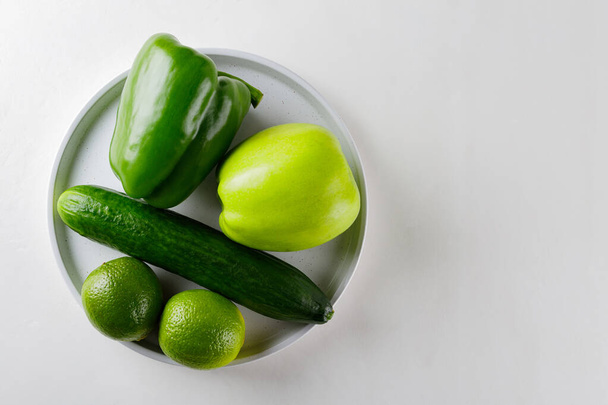 Various green vegetables and fruits on a white plate. Green apple, cucumber, bell pepper and lime on a white table. Copy space. Flat lay - Foto, Imagem