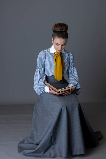 Una mujer de clase trabajadora eduardiana con una blusa de algodón rayado con un cravat amarillo mostaza, y una falda gris caminando sentada y leyendo sobre un fondo de estudio - Foto, imagen