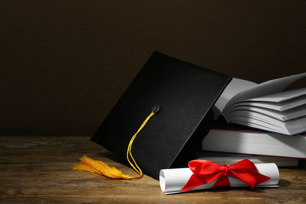 Graduation hat, books and diploma on wooden table against brown background, space for text - Fotó, kép