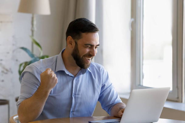 Счастливый молодой человек празднует успех интернета в домашнем офисе. - Фото, изображение