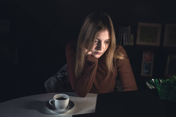 Μια νεαρή γυναίκα εργάζεται πίσω από ένα φορητό υπολογιστή τη νύχτα με ένα φως λαμπτήρα. - Φωτογραφία, εικόνα