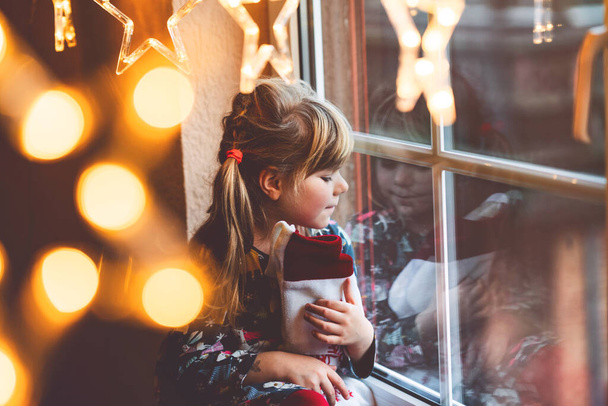 Malá školačka drží šálek Santa Claus bota s dárkem s názvem Nikolausstiefel v němčině. Šťastné dítě čeká na dovolenou u okna s vánoční světla v zimě. Útulná rodinná oslava Vánoc. - Fotografie, Obrázek