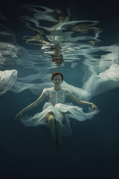 A girl in a white dress posing underwater on a dark background as if she were in zero gravity - Photo, image
