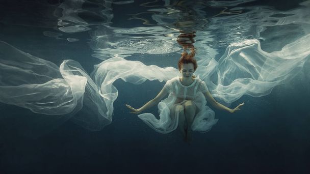 A girl in a white dress posing underwater on a dark background as if she were in zero gravity - Foto, Bild