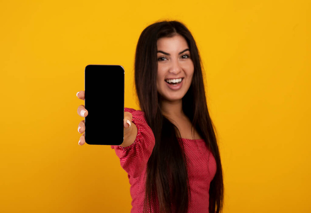 Mockup для застосунку або веб-сайту. Вірменська жінка тримає смартфон з чистим екраном, жовтий фон студії - Фото, зображення