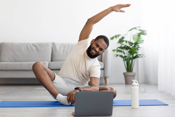 Happy young black bearded athlete man in white clothes do stretching or exercises for hands on mat - Photo, Image
