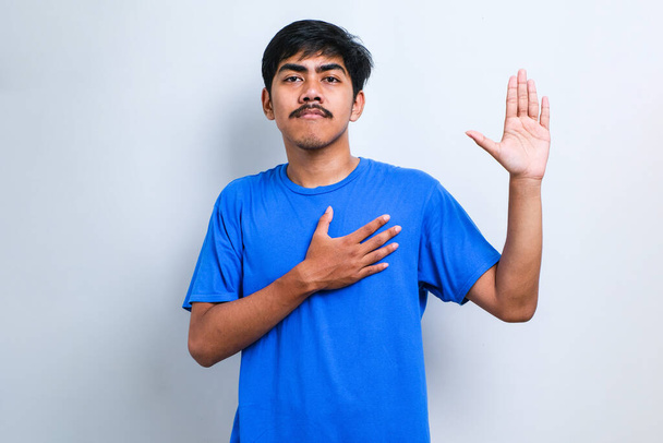 Junger gutaussehender asiatischer Mann in lässigem T-Shirt vor weißem Hintergrund lächelnd fluchend mit Hand auf Brust und erhobenen Fingern, Treueschwur - Foto, Bild