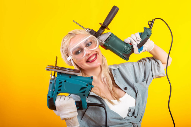 Beautiful girl with an electric drill and an electric jigsaw. Yellow background. - Valokuva, kuva