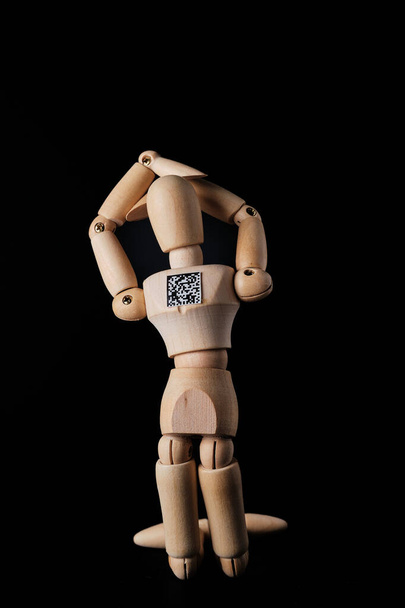 A wooden mannequin with a QR code is kneeling on a dark background. Selective focus. The concept of the trend of assigning QR codes to people in connection with countering the new Covid virus infection. A minimalist metaphor is the leash of global di - Fotoğraf, Görsel