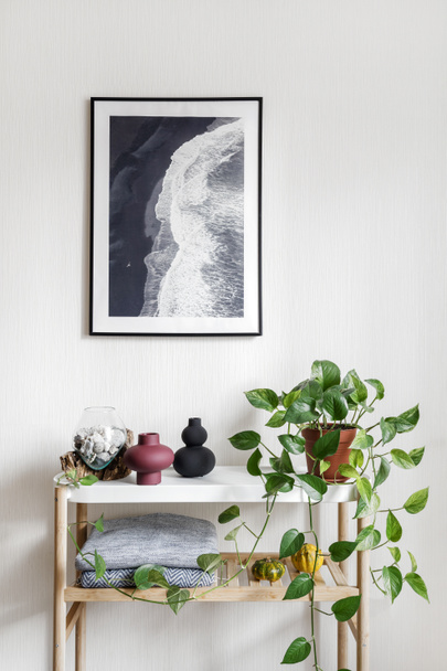 Picture frame with scenic ocean view hanging on wall over home decor and green potted plant. Vertical shot of vase with seashells standing on minimalistics white table in hygge apartment - Photo, image