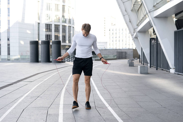 Concept of sport equipment. Full length view of good shaped sportsman training with skipping rope outdoors, warm up body before run on the street. Athlete preparing to cardio exercise in city - Photo, Image