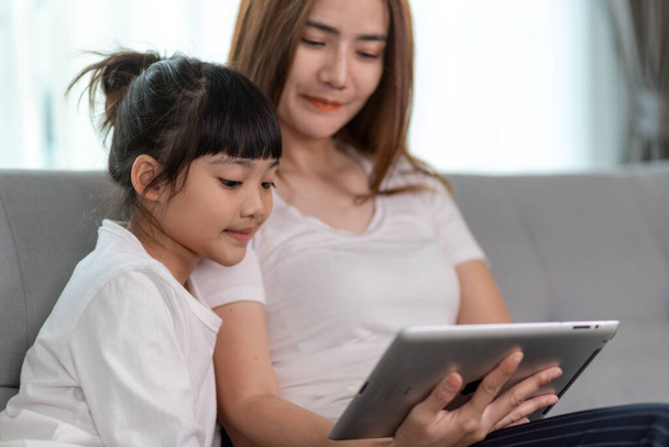 Beautiful Asian mother with daughter sitting on the sofa and playing video game on a tablet while spending time together at home - Zdjęcie, obraz