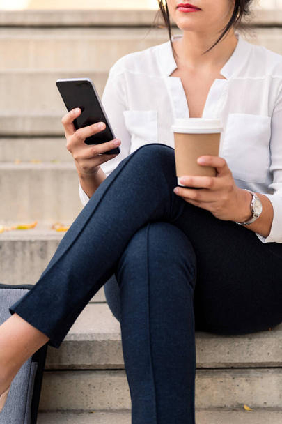 unrecognizable business woman consulting her phone sitting on a concrete staircase with a take away coffee in the other hand, concept of entrepreneur and urban lifestyle - Fotoğraf, Görsel