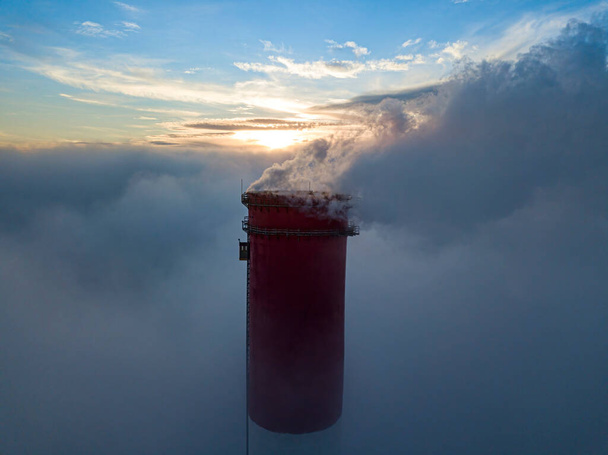 Wysoki komin elektrowni cieplnej nad mgłą w promieniach zachodu słońca. Widok drona z powietrza. - Zdjęcie, obraz
