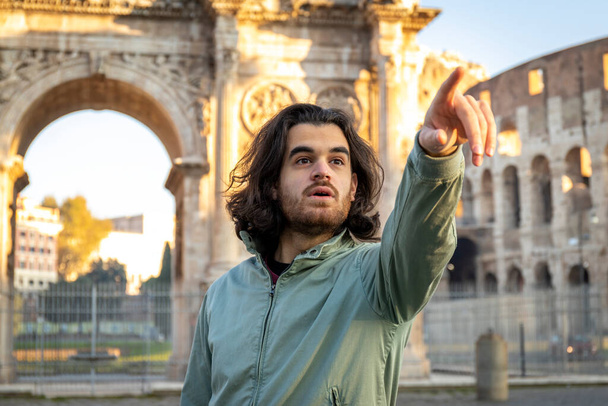 Hermoso hombre posa para una foto frente al Arco de Tito. El hombre con el pelo largo se sorprende al ver algo delante de él y señala a ella. - Foto, Imagen
