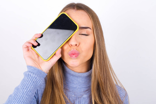 молодая женщина позирует со смартфоном в студии показывая экран за лицом - Фото, изображение