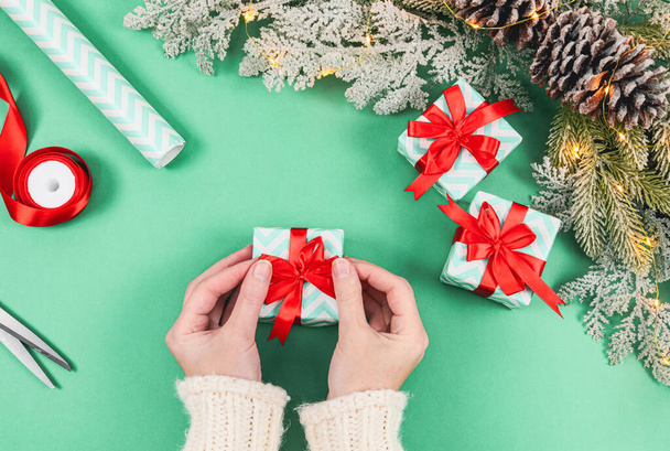 Females hands of a Caucasian woman in a white knitted sweater tie a red ribbon on a gift box, and next to scissors, paper and a spruce branch with a garland on a green background, top view, close-up. - Photo, Image