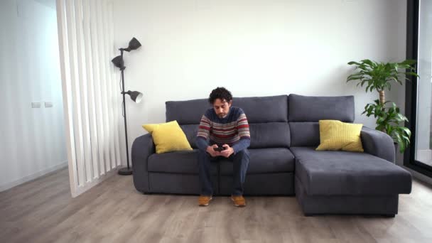 Homem bonito alegre usando seu telefone inteligente enquanto sentado no sofá em casa. Homem barbudo confiante está digitando no smartphone no sofá da casa. - Filmagem, Vídeo