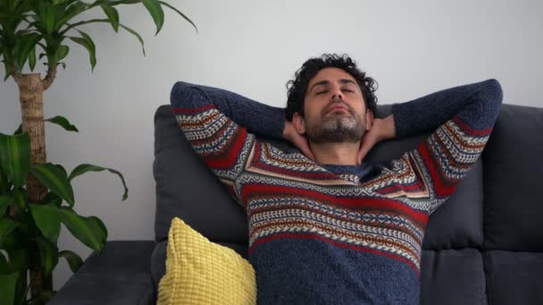 Όμορφος καυκάσιος άντρας που κρατάει τα μάτια κλειστά και κρατάει τα χέρια πίσω από το κεφάλι ενώ κάθεται στον καναπέ άνετα στο σπίτι. Απόλυτη χαλάρωση στον καναπέ στο σπίτι - Πλάνα, βίντεο