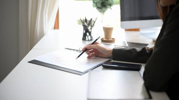 Zugeschnittenes Bild einer Managerin im schwarzen Anzug, die ihren Finanzbericht in einem Ordner am Schreibtisch ihres Büros überprüft. Stift zeigt auf Papierkram. - Foto, Bild