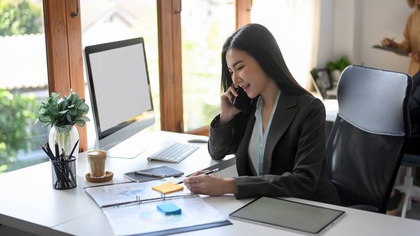 Впевнена азіатська бізнес-леді розмовляє по телефону зі своїм постачальником на її офісному столі. Жінка-менеджер має справу з партнером компанії по телефону
. - Фото, зображення