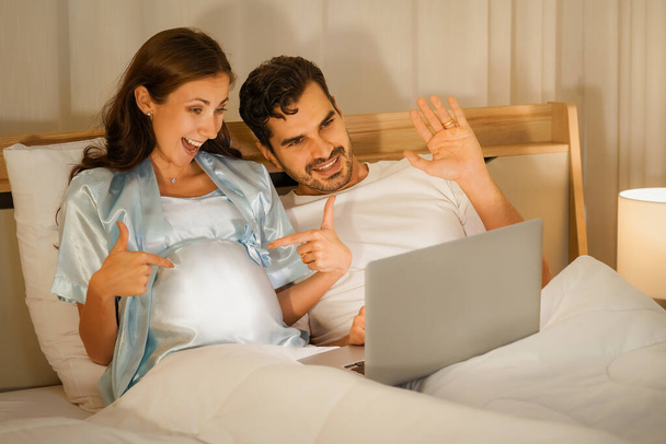 portrait of happy pregnant mother and husband having video chatting with friends on bed in bedroom - Foto, Imagen