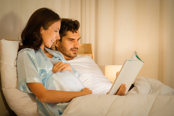 portrait of happy pregnant woman together with husband reading book on bed in bedroom - Foto, imagen