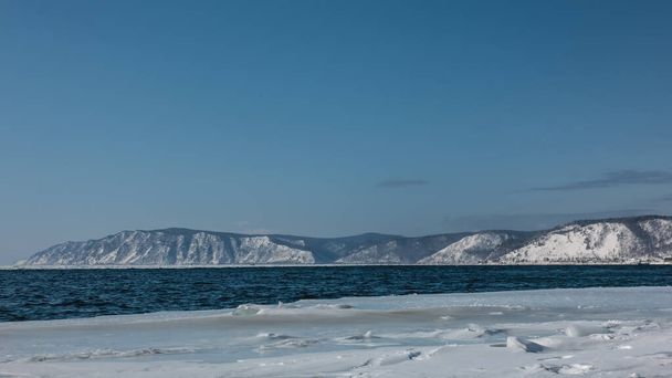 Υπάρχουν κυματισμοί στο σκούρο μπλε ποτάμι χωρίς πάγο. Χιόνι και πάγος στην ακτή. Μια δασώδης οροσειρά ενάντια σε έναν γαλανό ουρανό. Σιβηρία. Ποταμός Angara - Φωτογραφία, εικόνα