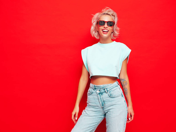 Young beautiful smiling female in trendy summer clothes. Sexy carefree woman posing near red wall in studio. Positive blond model having fun and going crazy. Cheerful and happy. In sunglasses - 写真・画像