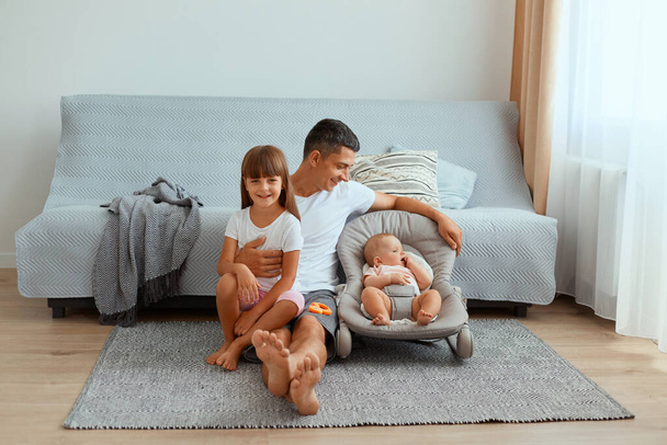 Image of brunette young father with his daughters sitting on floor near gray sofa, looking ta his infant child in rocking chair, spending time with children during weekend. - Foto, immagini