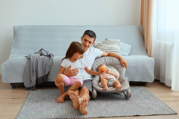 Horizontal shot of positive optimistic young father with his daughters sitting on floor near gray sofa, spending time with his children, expressing happiness, being glad to play with his kids. - Fotó, kép