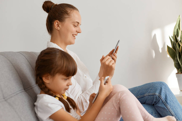 Side view portrait of smiling woman wearing white shirt and jeans sitting on sofa with her child, using cell phone for checking social networks or e-mail, her daughter playing game. - Foto, Imagem