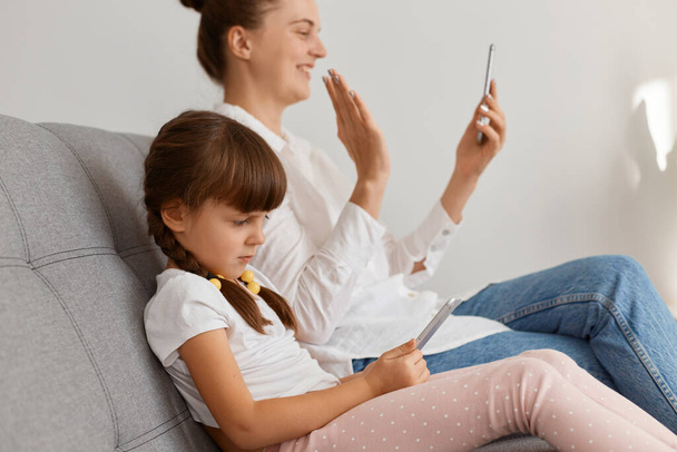 Side view portrait of positive attractive woman wearing white shirt and jeans sitting on sofa with her child, having video call or livestream, waving hand to followers. - Photo, image