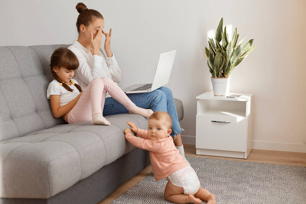 Indoor shot of cute toddler baby crawling to mother wearing shirt and jeans sitting on sofa with elder daughter, working on laptop, freelancer doing her work, being tire, feels eyes hurt, rubbing eye. - Φωτογραφία, εικόνα