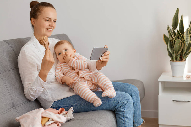 Side view portrait of smiling female with bun hairstyle wearing white shirt and jeans sitting on cough with her toddler kid, having video call or livestream, showing eco wooden toy to followers. - Foto, Imagem