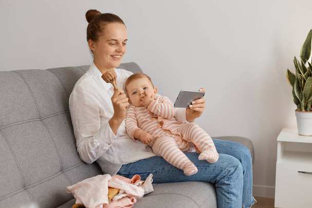 Smiling positive woman with hair bun wearing shirt and jeans sitting on sofa with her infant daughter and broadcasting livestream, showing wooden toy to followers, having video call via cell phone. - Photo, image