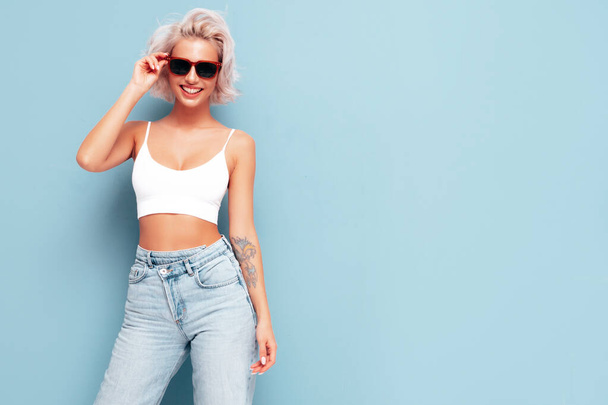Young beautiful smiling female in trendy summer clothes. Sexy carefree woman posing near blue wall in studio. Positive blond model having fun and going crazy. In sunglasses - Φωτογραφία, εικόνα
