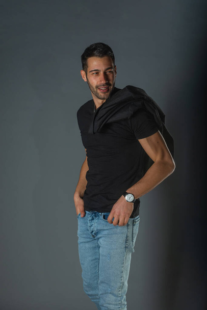 Forte modelo masculino muscular em jeans com t-shirt preta segurando seu casaco sobre os ombros - Foto, Imagem