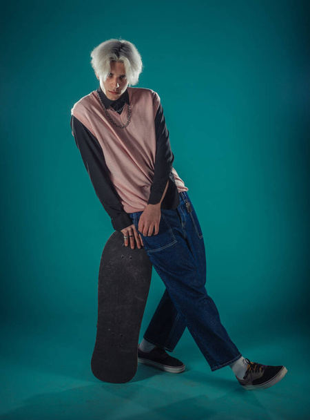 Young adult man in casual outfit wear looking at camera, holding skateboard near face and standing at studio on blue background - Fotografie, Obrázek