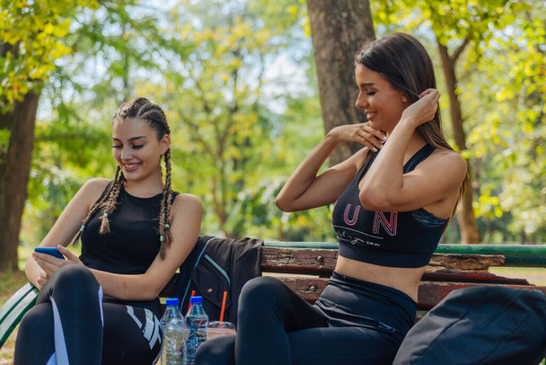 Hete aantrekkelijke vrouwen glimlachen en vegen op hun telefoons terwijl ze in een park zitten en een pauze nemen van hun sportieve activiteiten. - Foto, afbeelding