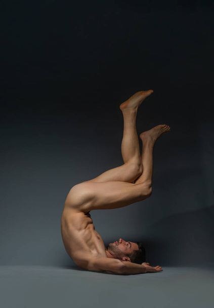 Upside down photo of a young naked man lying down on the floor in a dark studio with his legs up in the air - Zdjęcie, obraz